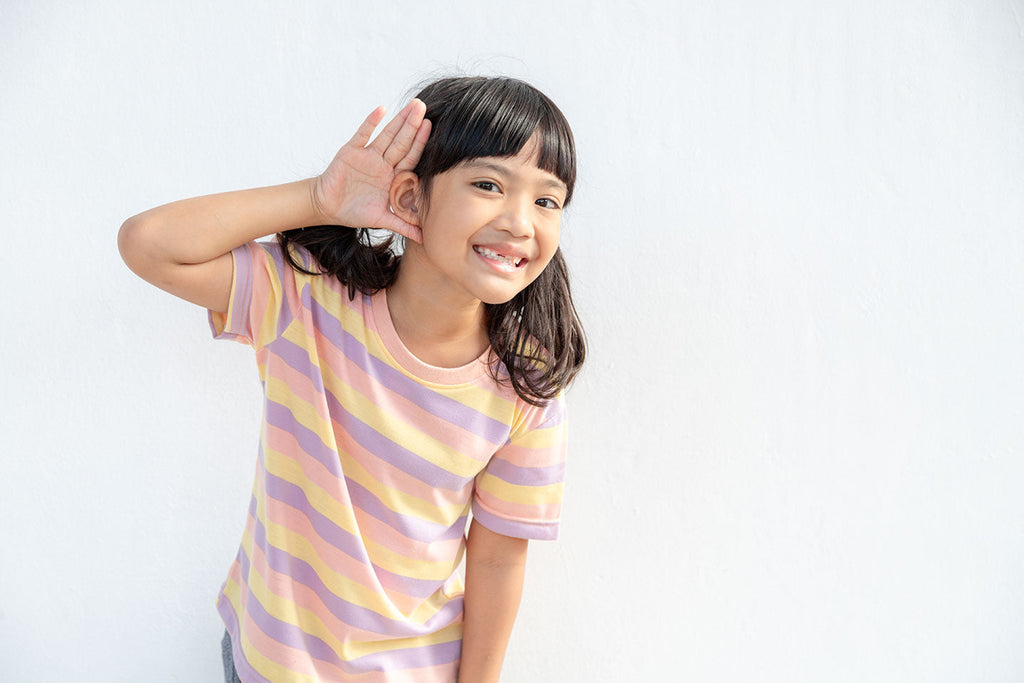 Smiling little girl holds her hand over her ears in an outward motion, implying that she is listening.