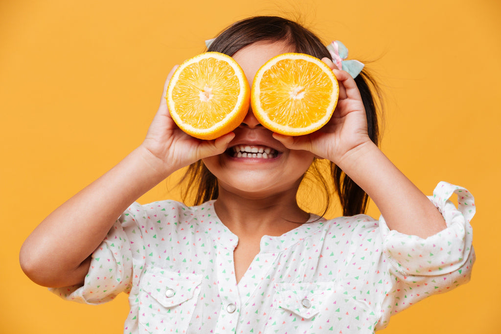 Little girl with Autism standing and smiling with 2 cut oranges in front of her eyes.