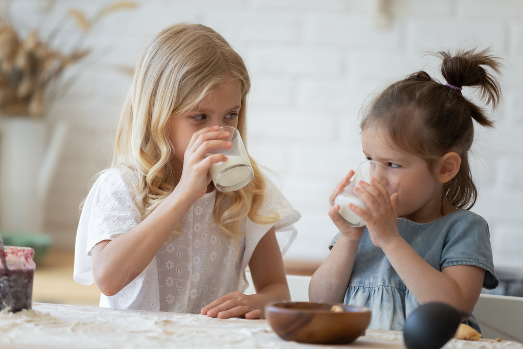 Two little sisters drinking morning glasses of milk with added probiotics for Autism spectrum disorder at a flour-covered kitchen table.